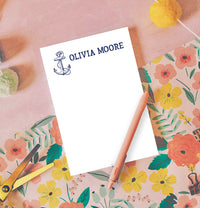 A nautical-themed notepad with an anchor design and the name 'Olivia Moore' printed at the top, accompanied by a pink pen and placed on a floral-patterned surface.