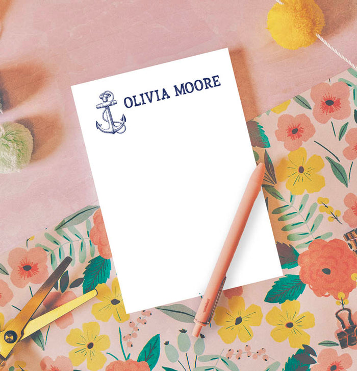 A nautical-themed notepad with an anchor design and the name 'Olivia Moore' printed at the top, accompanied by a pink pen and placed on a floral-patterned surface.