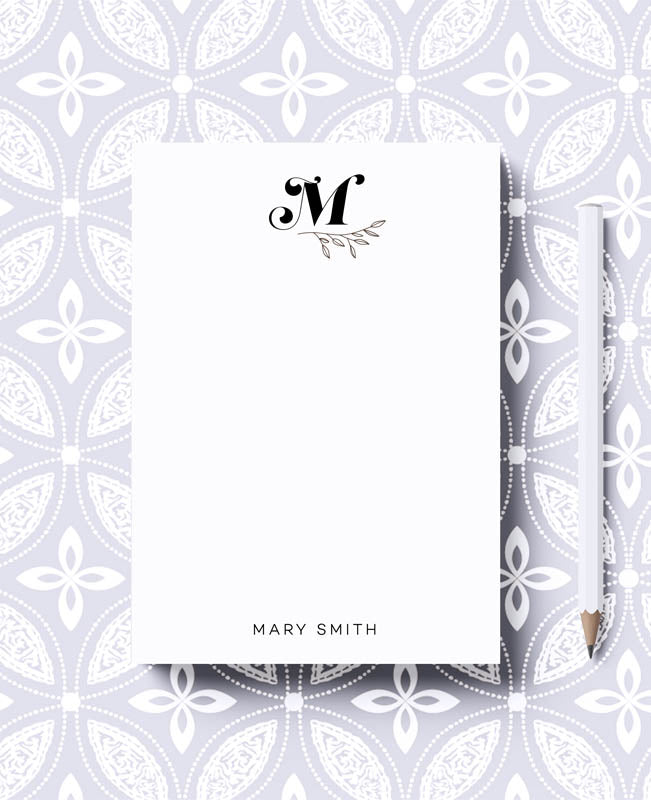 A personalized notepad with a bold monogram leaves design, featuring the initial 'M' and the name 'Mary Smith' on the cover. The notepad is presented on a background with a geometric pattern.