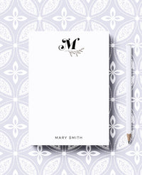 A personalized notepad with a bold monogram leaves design, featuring the initial 'M' and the name 'Mary Smith' on the cover. The notepad is presented on a background with a geometric pattern.