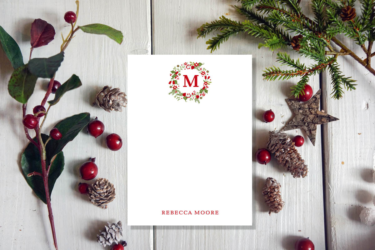 A white notepad with a Christmas garland design, placed on a wooden surface surrounded by pine cones and red berries.
