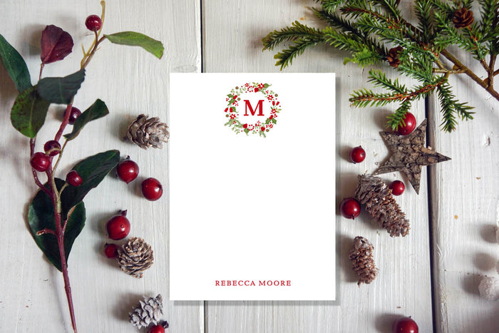 A white notepad with a Christmas garland design, placed on a wooden surface surrounded by pine cones and red berries.