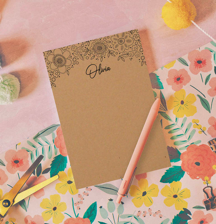 A floral brown kraft paper notepad with a customized name 'Diana' printed on the front, accompanied by a pink pen and placed on a table with a patterned background.