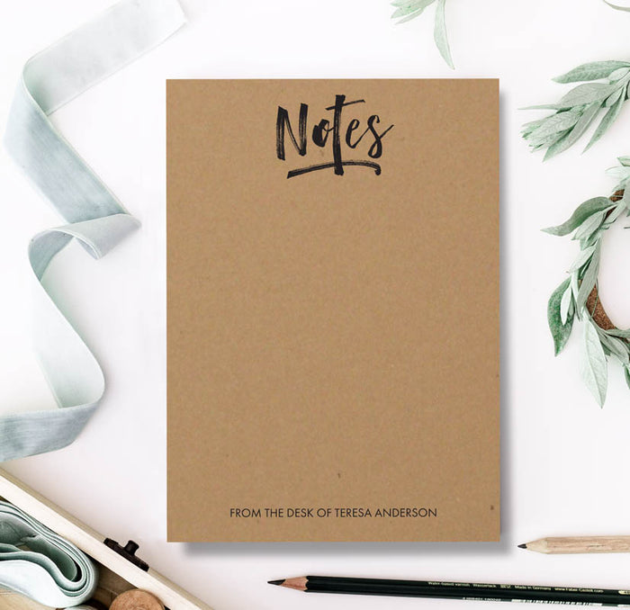A brown kraft paper notepad with the word 'Notes' written on the cover, placed on a desk with a pen and inkwell beside it.