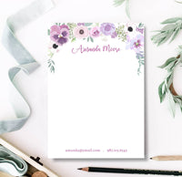 A personalized notepad with a floral design at the top, including purple and pink flowers and a custom name in cursive script. The notepad is placed on a desk with stationery supplies around it.