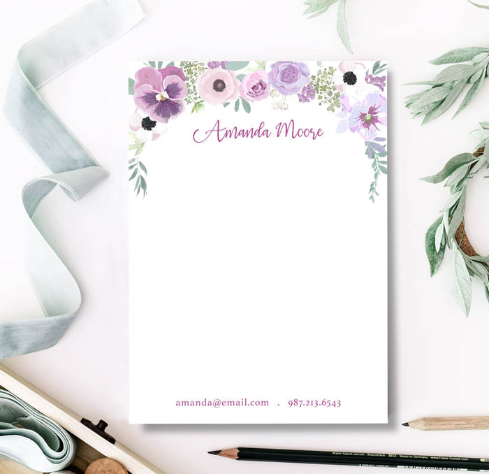 A personalized notepad with a floral design at the top, including purple and pink flowers and a custom name in cursive script. The notepad is placed on a desk with stationery supplies around it.